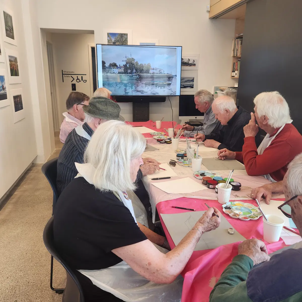 People seated around a table taking part in a watercolour art class. Paint palettes, brushes and paper are spread across a table covered with pink protective cloths, while a large screen at the front displays a landscape painting being used as a reference. The class is held in a gallery-like space with framed artwork on the walls.