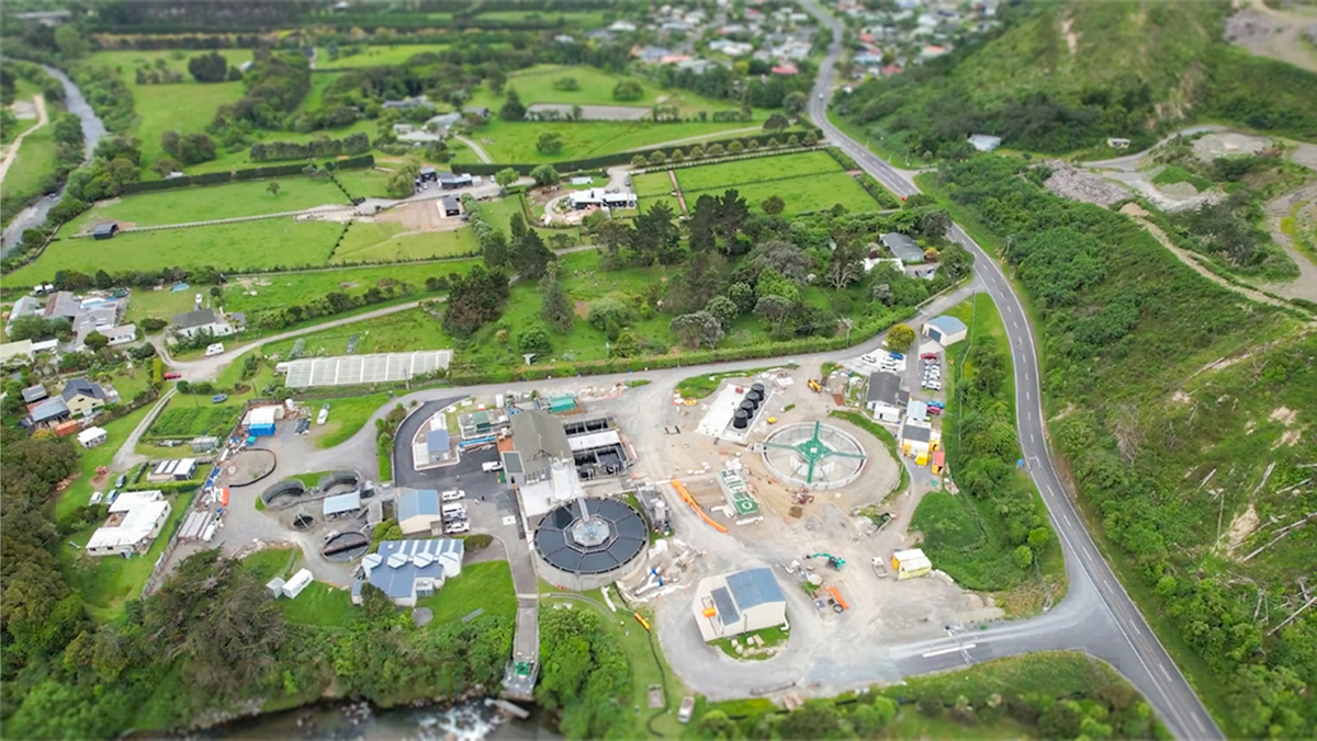 Aerial view of a water treatment plant showing circular treatment tanks, buildings, construction areas, and access roads, surrounded by green fields, trees, nearby homes, and a winding road along a hillside.