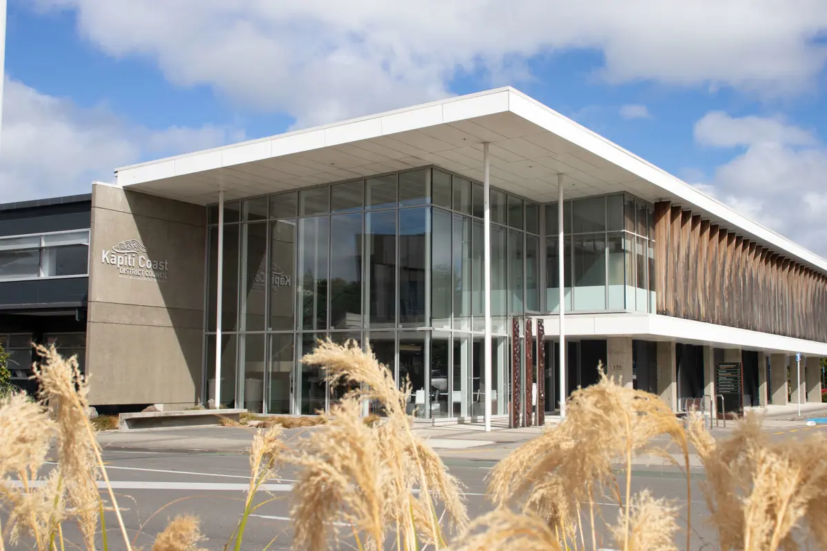 Modern civic building with a wide, flat white roof and large glass façade, supported by slim columns, viewed from a low angle. Tall golden grasses fill the foreground, with a mostly empty paved area in front and a partly cloudy sky above; “Kāpiti Coast District Council” signage is faintly visible on the left side of the building.