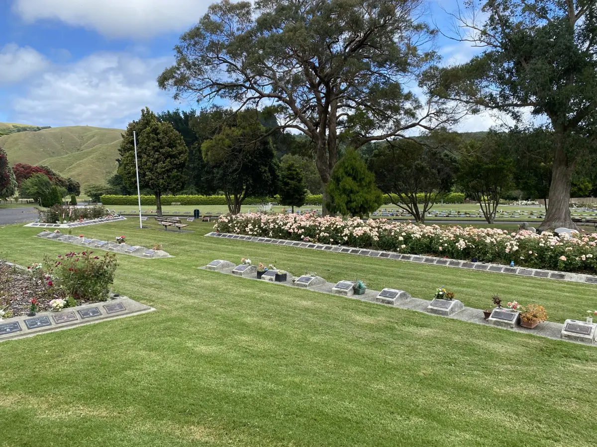 Well-maintained cemetery with rows of plaques and flower beds and large trees set against rolling green hills and a partly cloudy sky.