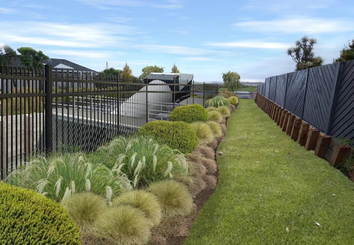 Landscaped garden with grasses and shrubs running alongside a fenced pump station structure, with a grass pathway and boundary fence on the right under a blue sky.