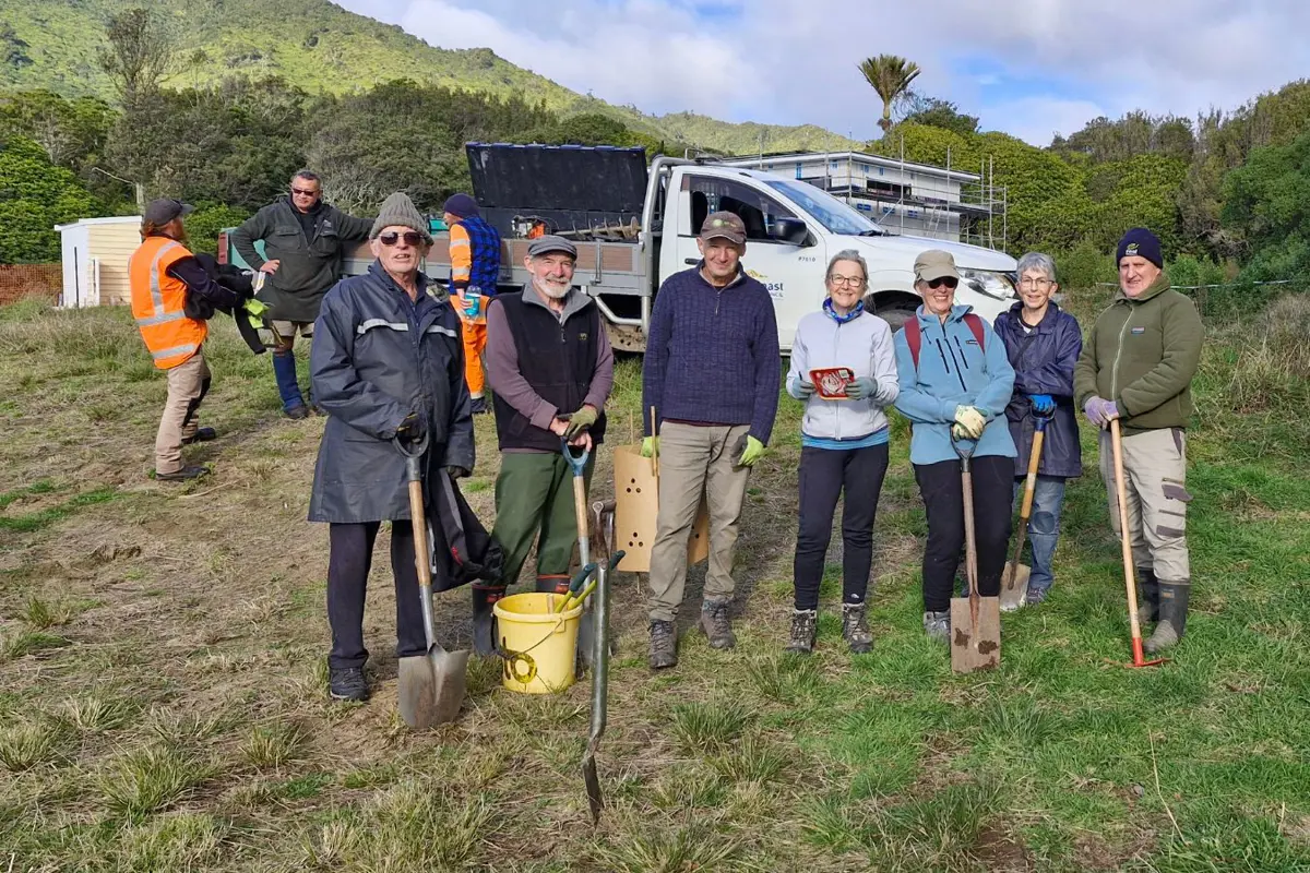 A group of people holding shovels stand in front of a truck with a mountain and native bush in the background.