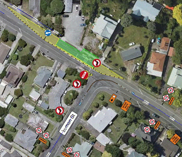 Aerial traffic management plan showing a residential intersection with temporary road layout changes. The main road runs diagonally across the image, with a side street joining from the lower centre. Green-highlighted road markings indicate a temporary work area near the intersection. Multiple traffic control signs, including "No Entry", directional arrows, and speed restriction signs, are positioned around the site. Orange barriers and temporary traffic management equipment are placed on the eastern side of the intersection. Nearby houses, driveways, parked vehicles, and trees are visible surrounding the work area.